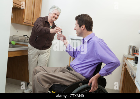 Home aiuto alla salute offrendo un bicchiere di acqua ad un uomo in carrozzella con lesioni del midollo spinale Foto Stock