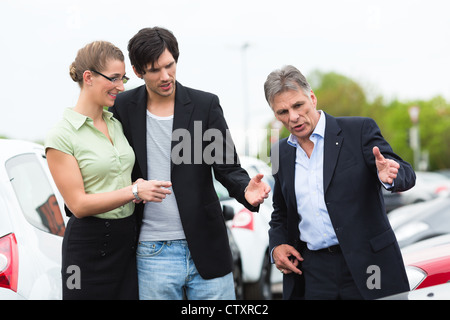 Coppia rivenditore di auto e coppia giovane in piedi sul posto di parcheggio in concessionaria nella parte anteriore delle vetture Foto Stock