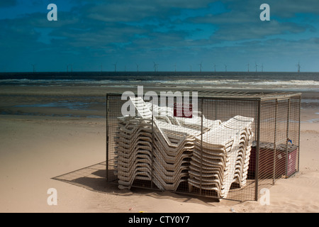 Sedie a sdraio impilati e recintata su una spiaggia deserta con turbine eoliche in background Foto Stock