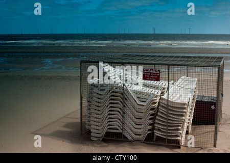 Sedie a sdraio impilati e recintata su una spiaggia deserta con turbine eoliche in background Foto Stock