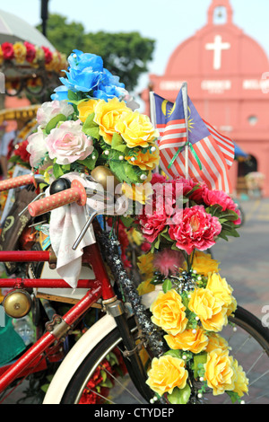 Coloratissimo risciò bicicletta ricoperto di fiori di plastica nella parte anteriore della storica Chiesa di Cristo in Malacca. Foto Stock