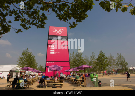 Spettatori riposo a tavoli e panche sotto una grande e accogliente segno gateway nel Parco Olimpico durante le Olimpiadi di Londra 2012. Questa terra è stata trasformata in un 2.5 sq km complesso sportivo, una volta che le imprese industriali ed ora sede di otto luoghi di interesse tra cui il principale arena, Aquatics Centre e Velodromo plus degli atleti del Villaggio Olimpico. Dopo le Olimpiadi, il parco è noto come Queen Elizabeth Olympic Park. Foto Stock