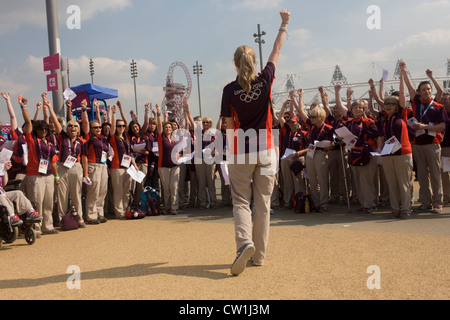 Con lo stadio principale e orbita arte dietro la torre, un coro di volontariato creatori di giochi di cantare per il divertimento di spettatori in arrivo nel parco olimpico durante le Olimpiadi di Londra 2012. Uno di loro numero pubblicizzato in un Olympic newsletter per i cantanti di unirsi nella risultante in 100 chiedono di aderire. I volontari sono chiamati 'Giochi Makers', come stanno contribuendo a rendere i giochi accadere. (Più didascalie in Descrizione.). Foto Stock