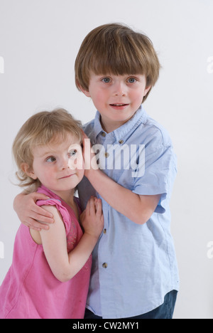 Ragazzo giovane trattiene Baby sorella, che copre le sue orecchie cercando avidamente verso la telecamera, ritratto in studio isolato su bianco Foto Stock