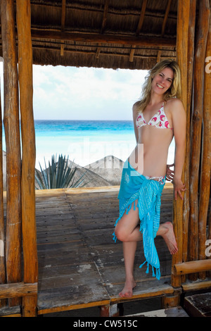 Messico, Cancun, giovane donna bionda appoggiata contro la porta della capanna Palapa e sorridente Foto Stock
