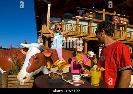 Famiglia giovane nella parte anteriore del mountain resort hotel, genitori seduti ad un tavolo del bar, giovane figlia in una vita di plastica di dimensioni mucca, Francia, Savoie, Alpi Foto Stock
