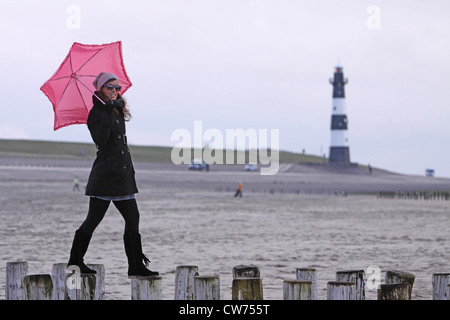 Giovane donna con ombrello rosa in equilibrio su sperone dighe con il faro in background, Paesi Bassi Zeeland, Breskens Foto Stock