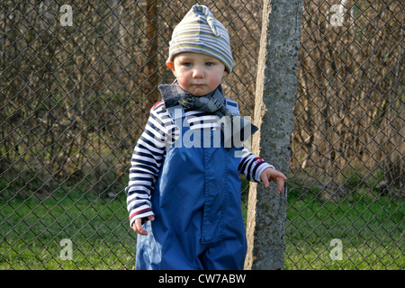 Little Boy nascita beanie e impermeabile dungaree permanente al giardino recinto, Germania Foto Stock