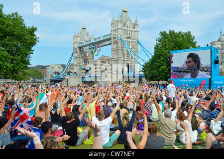 La gente della folla siede sull'erba vista i giochi olimpici di Londra 2012 Il grande schermo della TV mostra le mani in su e il Ponte della Torre ondeggiante Potters Fields Park Southwark Inghilterra Regno Unito Foto Stock