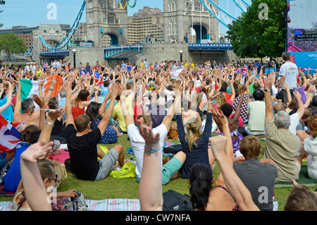 La gente della folla siede sull'erba vista i giochi olimpici di Londra 2012 Il grande schermo della TV mostra le mani in su e il Ponte della Torre ondeggiante Potters Fields Park Southwark Inghilterra Regno Unito Foto Stock