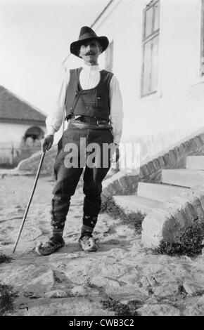 Il serbo uomo che indossa hat, giubbotto, belted pantaloni e scarpe a punta con canna in piedi al di fuori del palazzo in Iugoslavia, circa Foto Stock