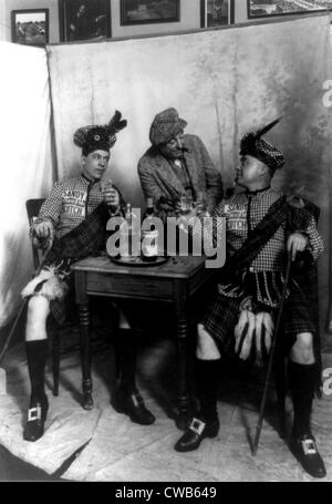 Due uomini che indossano kilts seduto al tavolino, bere Sandy MacDonald scotch, con uomo in piedi e appoggiata contro il tavolo, Foto Stock