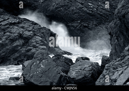 Foto in bianco e nero di onde che si infrangono attraverso un foro di sfiato nel shorline rocciose di Kynance Cove in Cornovaglia Foto Stock