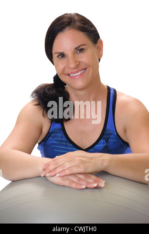 Ritratto di una bella bruna donna nella sua 40s blu da indossare abbigliamento allenamento, sorridente e appoggiata su un livello di grigio palla ginnica. Foto Stock
