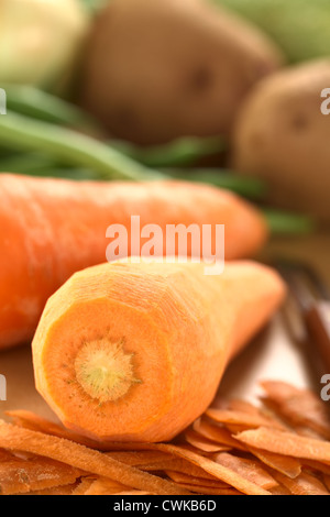 Materie sbucciate la carota con sbucciatore e altri ortaggi o legumi (fagioli verdi e patate) sulla tavola di legno (messa a fuoco selettiva) Foto Stock