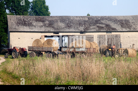 Agricoltore prelievo rotonda di grandi dimensioni balle di fieno con il suo trattore per il rimessaggio in Fattoria Barn. Zawady Polonia centrale Foto Stock