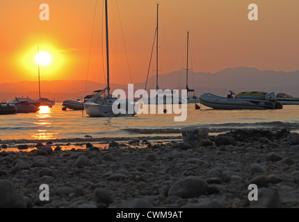 Spiaggia sassosa, il Lago di Garda al tramonto, Camping Lido, Pacengo, Italia, con barche ormeggiate in background Foto Stock