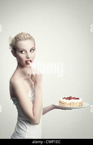 Giovane donna tenendo la torta di fragole, leccare la crema off dito, ritratto Foto Stock