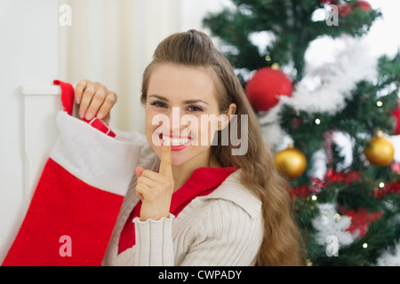 Sorridente giovane donna mettere in regalo calze di Natale e mostra il gesto di shh Foto Stock