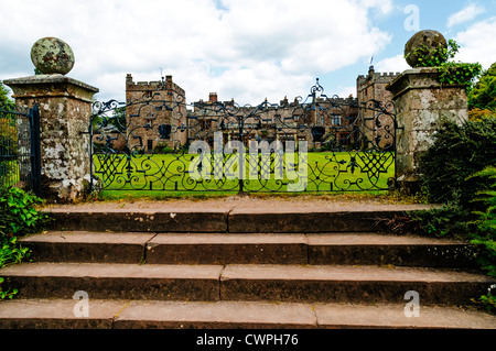 Il prato e la facciata del castello di Muncaster con due merlate torri quadrate visto attraverso il pattern dei cancelli in ferro battuto Foto Stock