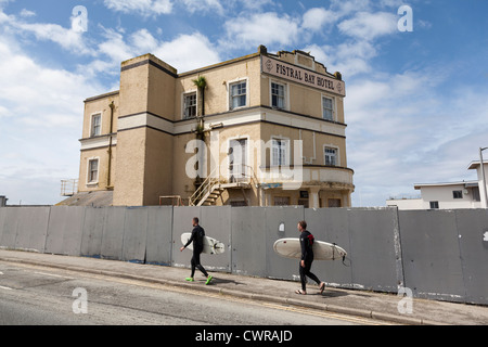 Surfer con tavole andare passato abbandonato Fistral Bay Hotel, Newquay, Cornwall, Regno Unito. Foto Stock