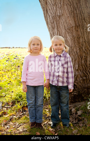 Twin bionda ragazze sono in piedi accanto a ogni altro di fronte a un albero. Foto Stock