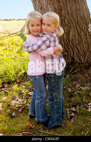 Twin bionda ragazze sono in piedi accanto a ogni altro di fronte a un albero. Foto Stock