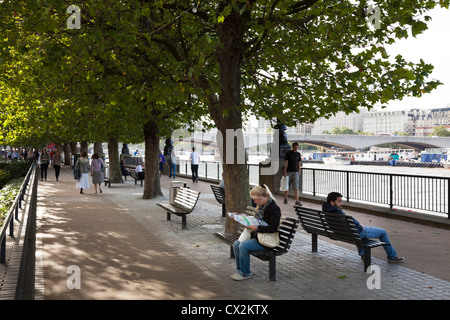 La gente seduta sulle panche sotto l'ombra di alberi sulla riva sud del Tamigi vicino a Waterloo bridge Foto Stock