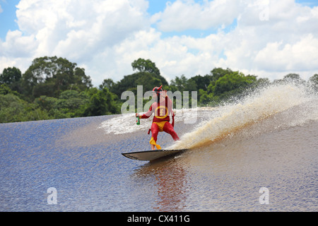 Uomo Barbuto in costume da supereroe navigare un'onda tenendo in mano una bottiglia di birra sul fiume Kampar a Sumatra, Indonesia Foto Stock