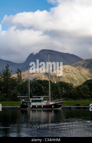 Uno yacht è ormeggiato sulle rive del Caledonian Canal come Ben Nevis, la montagna più alta della Gran Bretagna, torri su di esso in Foto Stock