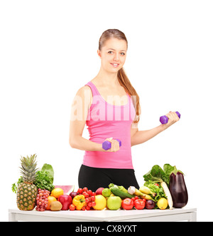 Donna sorridente tenendo pesi e tavolo pieno di frutta e verdura isolati su sfondo bianco Foto Stock