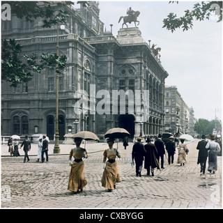 Vista colorata dei pedoni che camminano lungo l'Opernring di fronte al Teatro dell'Opera di Vienna, Vienna, Austria, 1907. Alcune delle donne tengono gli ombrelli sopra le loro teste. Sulla facciata sono visibili le statue di Ernst Julius Hähnel di Erato e di Harmony su cavalli alati. (Foto di Burton Holmes) Foto Stock