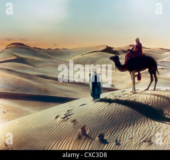 Foto colorata di una persona che cavalcava un cammello fermata su una duna di sabbia nel deserto al tramonto, Giza, Egitto, 1906. Un uomo, forse una guida, si trova di fronte al cammello e tiene una frusta sulla schiena. (Foto di Burton Holmes) Foto Stock