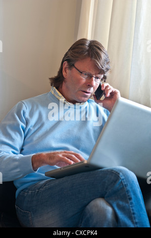 Uomo maturo a casa controllo schermo informazioni sul suo computer portatile e parlando su un iPhone 4s smartphone Foto Stock