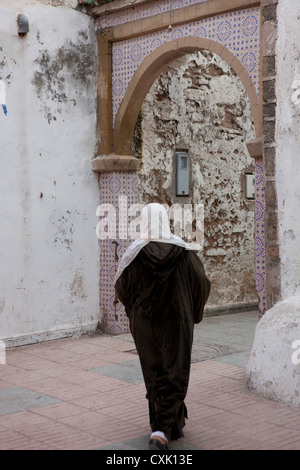 Local donna marocchina in djellaba e testa sciarpa a camminare verso un arco modo Essaouira, Marocco Foto Stock