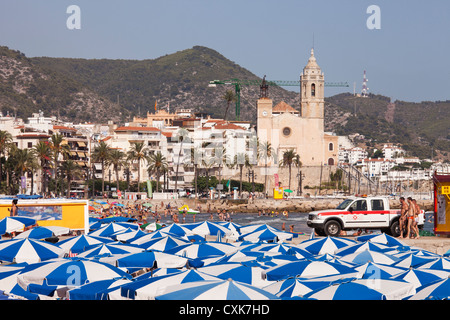 Guardando attraverso la spiaggia di Sitges, Spagna, uno può vedere il blue-e-ombrelloni bianchi e molti frequentatori di spiaggia. Foto Stock