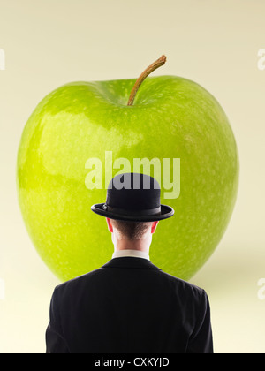 Uomo con vestito nero e cappello da bowling di fronte a una grande mela verde. Basato sul pittore surreale Rene Magritte. Foto Stock