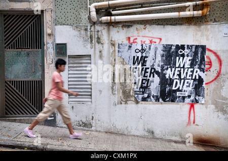 Una donna cammina passato manifesti stracciati e graffiti sul lato di un edificio residenziale in Sheung Wan, isola di Hong kong Foto Stock