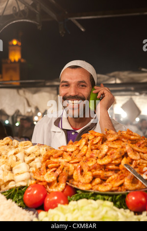 Cibo locale venditore sorridente Piazza Jamaa El Fna a Marrakech con minareto della moschea di Koutoubia in background, Marocco Foto Stock