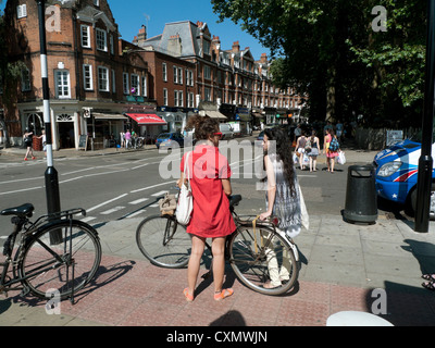 Giovani donne amiche in abiti estivi con biciclette in piedi Parlando all'angolo di East Heath Road in Hampstead North LONDRA NW3 REGNO UNITO KATHY DEWITT Foto Stock