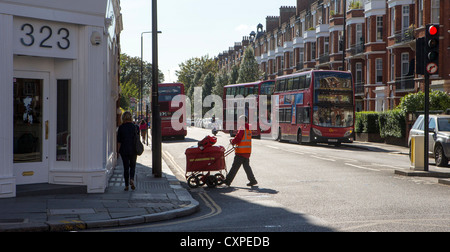 Un postino Londra attraversando la strada e wheeling il suo email carrello / carrello di fronte a lui, con autobus rossi di Londra dietro. Foto Stock