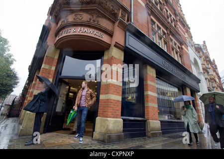 Gli amanti dello shopping con ombrelloni passano davanti all'outlet di moda Gieves & Hawkes a Kings Road, Chelsea, Londra, Regno Unito, catturando il patrimonio sartoriale britannico, il negozio di abbigliamento maschile di lusso e la vita di strada di tutti i giorni in condizioni di pioggia in un'elegante strada di Londra Foto Stock