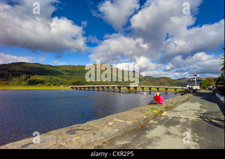 Adulto maschio femmina rilassatevi sulla mawddach riverside, bianco in legno ponte a pedaggio e il vecchio segnale ferroviario in background, albero colline coperte Foto Stock