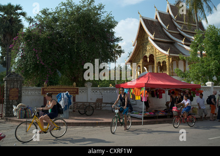 Ciclo di turisti passato il tempio buddista nella motivazione del Royal Palace, a Luang Prabang, Laos Foto Stock