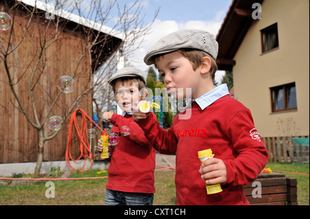 Twin boys, 4, indossando tappi piatti e soffiare bolle di sapone in un giardino Foto Stock