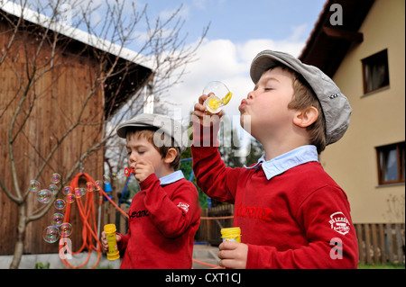 Twin boys, 4, indossando tappi piatti e soffiare bolle di sapone in un giardino Foto Stock