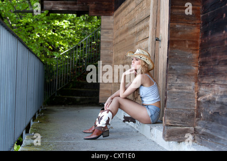 Giovane donna in posa mentre si è seduti indossando il denim hot pants, un cappello e un paio di stivali da cowboy, stile occidentale Foto Stock