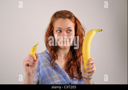 Giovane donna con i capelli rossi confrontando una molto grande e di una piccola banana nelle sue mani Foto Stock