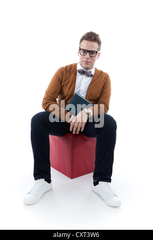 Giovane uomo con gli occhiali e un filtro bow tie in possesso di un libro, agenda, seduto su un cubo rosso Foto Stock