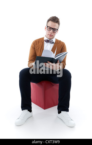 Giovane uomo con gli occhiali e un filtro bow tie sfogliando un libro, agenda, seduto su un cubo rosso Foto Stock